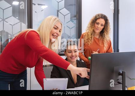 Drei lächelnde weibliche Profis haben sich aktiv mit einem Tablet und Monitor in einem eleganten Büro an einer gemeinsamen Aufgabe beteiligt Stockfoto