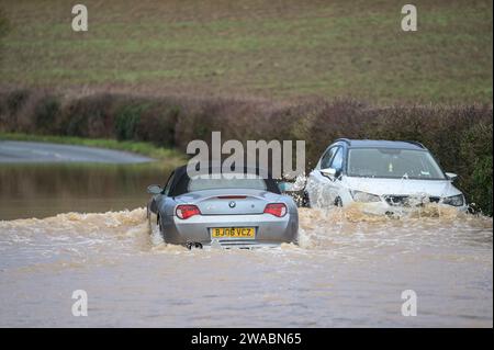 Offenham, Evesham, 3. Januar 2024 – Ein BMW-Fahrer versuchte und scheiterte, durch Hochwasser zu fahren, obwohl ein anderes Fahrzeug bereits auf derselben Strecke verlassen wurde. Der ältere Fahrer drehte seinen Motor auf, als er durchfuhr, aber seine Geschwindigkeit war zu hoch und Wasser stieg in seinen Motor ein. Das Fahrzeug ging schließlich wieder los, aber es kam nur wenige Meter vom Trockenland entfernt zu einer weiteren Haltestelle. Ein Straßenarbeiter kam dann an und legte als letzte Beleidigung ein Schild mit der Aufschrift „Road Closed“ vor sein Fahrzeug. Es geschah neben dem Offenham Caravan Park, der sich in der Nähe von Evesham in Worcestershire befindet, wo der Fluss Avon niederfließt Stockfoto