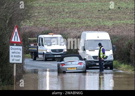 Offenham, Evesham, 3. Januar 2024 – Ein BMW-Fahrer versuchte und scheiterte, durch Hochwasser zu fahren, obwohl ein anderes Fahrzeug bereits auf derselben Strecke verlassen wurde. Der ältere Fahrer drehte seinen Motor auf, als er durchfuhr, aber seine Geschwindigkeit war zu hoch und Wasser stieg in seinen Motor ein. Das Fahrzeug ging schließlich wieder los, aber es kam nur wenige Meter vom Trockenland entfernt zu einer weiteren Haltestelle. Ein Straßenarbeiter kam dann an und legte als letzte Beleidigung ein Schild mit der Aufschrift „Road Closed“ vor sein Fahrzeug. Es geschah neben dem Offenham Caravan Park, der sich in der Nähe von Evesham in Worcestershire befindet, wo der Fluss Avon niederfließt Stockfoto