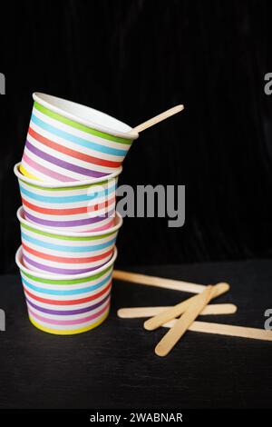 Ein Stapel bunt gestreifter Pappbecher und Holzrührer auf schwarzem Hintergrund. Die Cups sind mit horizontalen Streifen in verschiedenen hellen Farben verziert Stockfoto