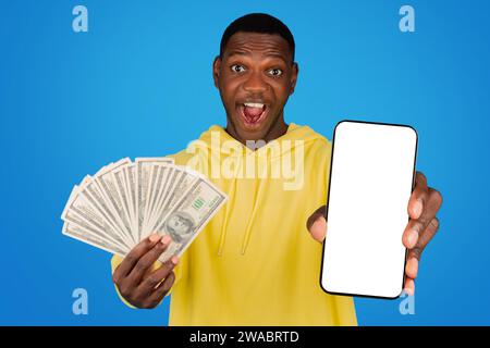 Ein aufgeregter junger Mann in einem gelben Hoodie zeigt einen Stapel Geld in einer Hand und einen leeren Bildschirm Stockfoto