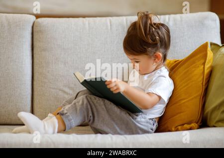 Süßes Kleines Mädchen, Das Sich Auf Der Couch Mit Interessantem Buch Entspannt Stockfoto