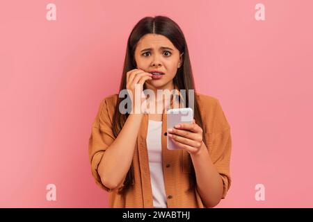 Ängstliches Teenager-Mädchen beißt sich die Nägel, während sie auf das Smartphone blickt. Stockfoto