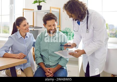 Ein freundlicher Gynäkologe berät Paare über die Schwangerschaft und zeigt ihnen das Clipboard mit Testergebnissen. Stockfoto