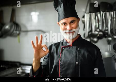 Reifer Koch im schwarzen Mantel, der das OK-Zeichen in seiner Küche macht Stockfoto