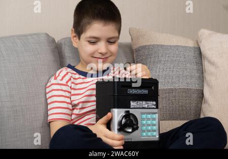 Ein kleiner Junge steckt eine Münze in eine sichere Schweinebank. Finanzkompetenz für Kinder. Stockfoto