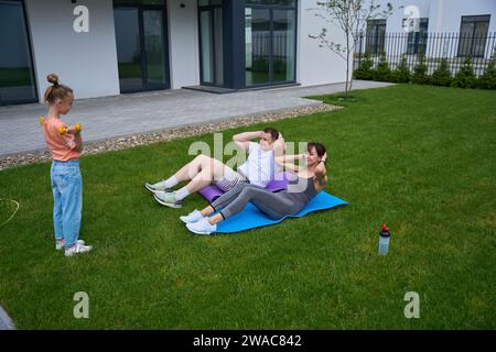 Mädchen mit Kurzhanteln, die ihre Mom und ihren Dad anschauen, wie sie auf dem grünen Hof knirschen Stockfoto