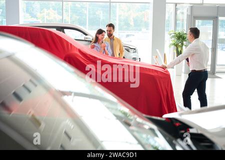 Der Manager des Autohändlers präsentiert das Fahrzeug unter roter Abdeckung Stockfoto