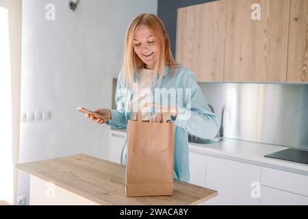 Online-Lieferung von Lebensmitteln zu Hause. Frau, die ihre Online-Bestellliste auf ihrem Handy überprüft. Eine junge Frau packt zu Hause Kisten mit Lebensmitteln aus Stockfoto