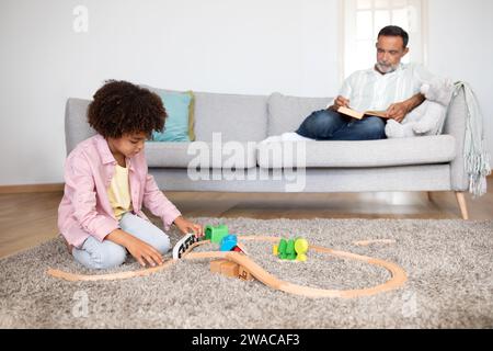 Enkel spielt beim Bauen der Spielzeugbahn, während der Großvater drinnen liest Stockfoto