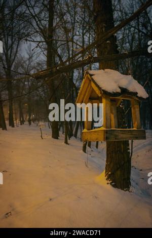 Vogelfutter im verschneiten Wald. Holzvogelhaus im Winterpark am Abend. Tierpflegekonzept. Handgefertigte Vogelfuttermittel im Schnee auf dem Baum. Stockfoto