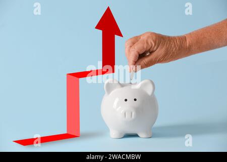 Hand einer Seniorin, die Münze in die Schweinebank steckt und einen Pfeil auf blauem Hintergrund erhöht. Konzept des steigenden Preises Stockfoto