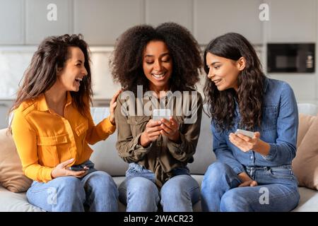 Drei multiethnische Freundinnen, die im Wohnzimmer auf Handys surfen Stockfoto