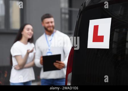 Fahrer und Kursleiter mit Klemmbrett in der Nähe des Autos im Freien, selektiver Fokus auf L-Platte. Fahrschule Stockfoto