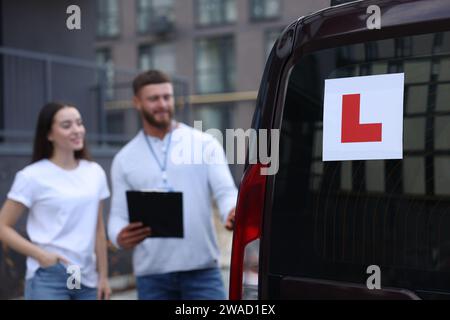 Fahrer und Kursleiter mit Klemmbrett in der Nähe des Autos im Freien, selektiver Fokus auf L-Platte. Fahrschule Stockfoto