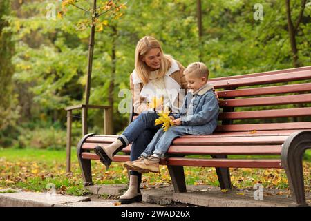 Glückliche Mutter und ihr Sohn verbringen Zeit zusammen mit trockenen Blättern auf Holzbank im Herbstpark Stockfoto