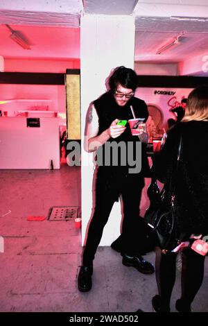 Mann in schwarzen, schwarzen Haaren gekleidet, schwarze Gläser mit einem Drink in einem roten Papierbecher, der ein lindgrünes Telefon auf einer Lagerparty hält, rosa Beleuchtung Stockfoto