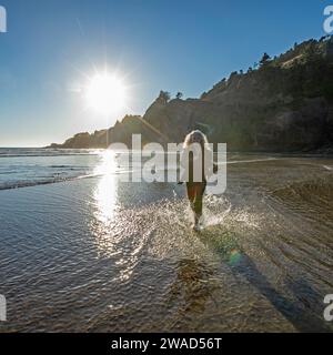 USA, Oregon, Newport, Frau, die am Sandstrand läuft und Wasser spritzt Stockfoto