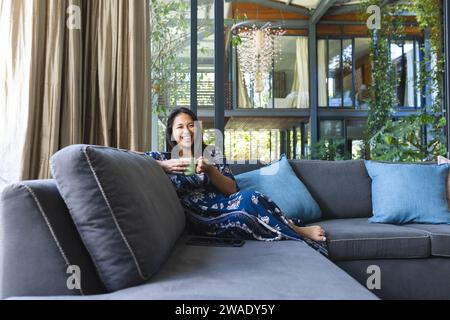 Porträt einer glücklichen asiatischen Frau auf Couch mit Kaffee zu Hause. Leben, Trinken und Lebensstil, unverändert. Stockfoto