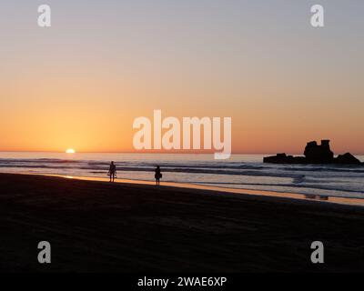 Am 3. Januar 2024 spazieren Sie zu zweit an einem Strand, während die Sonne am Horizont untergeht und ein Felsvorsprung direkt vor der Küste in Essaouira, Marokko, liegt Stockfoto