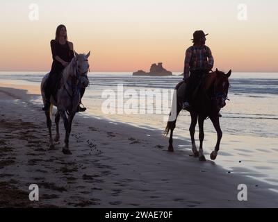 Männliche und weibliche Reiten an einem Sandstrand bei Sonnenuntergang mit einem Felsvorsprung dahinter und Licht reflektiert sich im Meer. Essaouira, Marokko, 3. Januar 2024 Stockfoto