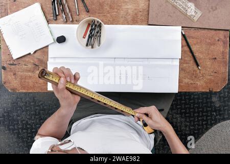 Draufsicht junger lateinischer Mann, der Messungen von handgemachter Holzflöte mit Arbeitstisch im Hintergrund macht Stockfoto