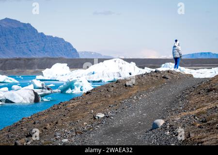 Ein junger Erwachsener steht an einer felsigen Küste in der Nähe eines Wassers, umgeben von schneebedeckten Bergen und einer Kulisse aus blauem Himmel Stockfoto