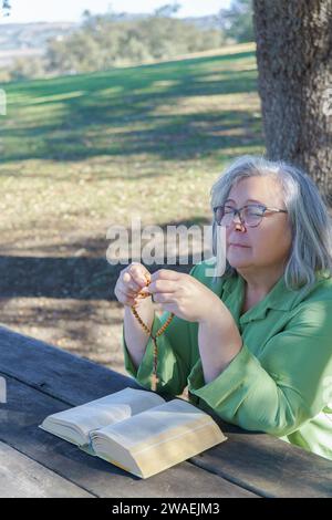 Ältere weißhaarige Frau mit Rosenkranzperlen in der Hand und einer Bibel, die mit geschlossenen Augen betet und an einem Holztisch im Wald sitzt. Stockfoto