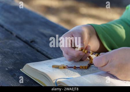 Von den Händen einer Frau, die die Rosenkranzperlen über einer Bibel beten, in der sonnigen Landschaft Stockfoto