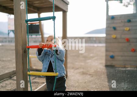 Das kleine Mädchen steht neben einer Seilleiter auf einem Spielplatz und schaut nach oben Stockfoto