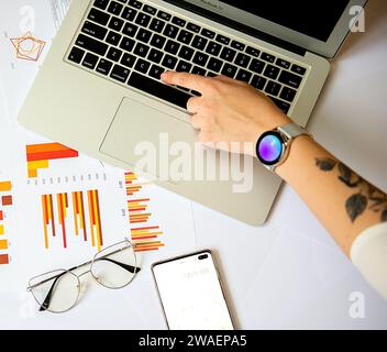 Eine Person, die auf einem Laptop an einem Schreibtisch mit Smartphone und Dokumenten schreibt Stockfoto