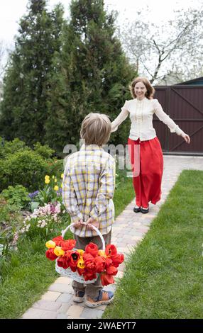 Der Junge steht und versteckt einen Korb mit Blumen hinter seinem Rücken, um eine Überraschung für einen Geburtstag oder Muttertag zu machen. Die Freude und das Glück der Mutterschaft. Selektiv Stockfoto