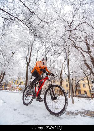 Ein Typ in einer orangen Jacke fährt mit dem Fahrrad durch eine Winterstadt zwischen schneebedeckten Bäumen. Aktiver Lebensstil im Winter Stockfoto