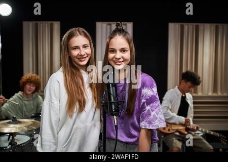 Lustige Teenager-Mädchen, die in die Kamera in der Nähe des Mikrofons schauen, während ihre Freunde Schlagzeug und Gitarre spielen Stockfoto