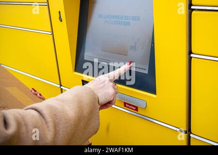 Innovative Paketlogistik - Nutzung der DHL Packstation 04.01.2024, Bad Camberg: Symbolfoto, Illustrationsbild, Symbolbild, Illustrationsfoto das Bild zeigt eine Frau im Anschnitt, die mit ihrem Smartphone über den Touchscreen einer DHL Paketstation interagiert, um ein Paket einzureichen. Sie ist konzentriert auf die digitale Schnittstelle fokussiert, während Ihre Hand das Mobiltelefon hält und die entsprechenden Eingaben auf dem Bildschirm der Paketstation tätigt um das Paket einlegen. Die Packstation, ein Paketautomatensystem von DHL, bietet seit 2001 in über 12,500 Standorten deutschlandwe Stockfoto