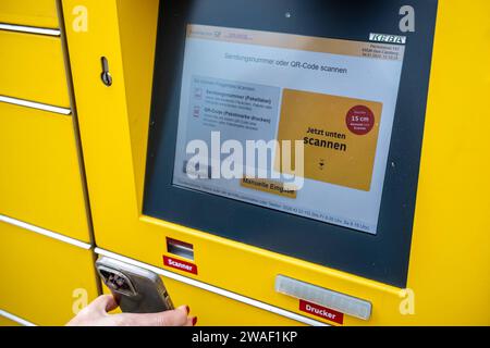 Innovative Paketlogistik - Nutzung der DHL Packstation 04.01.2024, Bad Camberg: Symbolfoto, Illustrationsbild, Symbolbild, Illustrationsfoto das Bild zeigt eine Frau im Anschnitt, die mit ihrem Smartphone über den Touchscreen einer DHL Paketstation interagiert, um ein Paket einzureichen. Sie ist konzentriert auf die digitale Schnittstelle fokussiert, während Ihre Hand das Mobiltelefon hält und die entsprechenden Eingaben auf dem Bildschirm der Paketstation tätigt um das Paket einlegen. Die Packstation, ein Paketautomatensystem von DHL, bietet seit 2001 in über 12,500 Standorten deutschlandwe Stockfoto