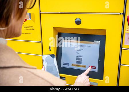 Innovative Paketlogistik - Nutzung der DHL Packstation 04.01.2024, Bad Camberg: Symbolfoto, Illustrationsbild, Symbolbild, Illustrationsfoto das Bild zeigt eine Frau im Anschnitt, die mit ihrem Smartphone über den Touchscreen einer DHL Paketstation interagiert, um ein Paket einzureichen. Sie ist konzentriert auf die digitale Schnittstelle fokussiert, während Ihre Hand das Mobiltelefon hält und die entsprechenden Eingaben auf dem Bildschirm der Paketstation tätigt um das Paket einlegen. Die Packstation, ein Paketautomatensystem von DHL, bietet seit 2001 in über 12,500 Standorten deutschlandwe Stockfoto