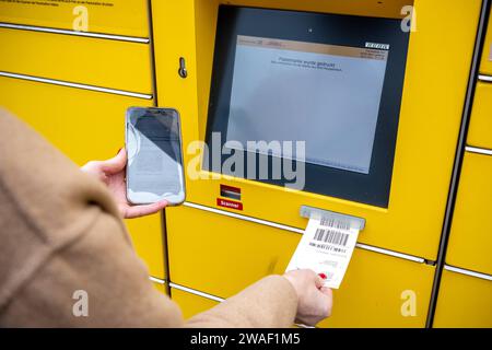 Innovative Paketlogistik - Nutzung der DHL Packstation 04.01.2024, Bad Camberg: Symbolfoto, Illustrationsbild, Symbolbild, Illustrationsfoto das Bild zeigt eine Frau im Anschnitt, die mit ihrem Smartphone über den Touchscreen einer DHL Paketstation interagiert, um ein Paket einzureichen. Sie ist konzentriert auf die digitale Schnittstelle fokussiert, während Ihre Hand das Mobiltelefon hält und die entsprechenden Eingaben auf dem Bildschirm der Paketstation tätigt um das Paket einlegen. Die Packstation, ein Paketautomatensystem von DHL, bietet seit 2001 in über 12,500 Standorten deutschlandwe Stockfoto