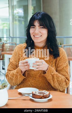 Porträt einer jungen lateinamerikanischen venezolanischen Frau, mit lockigen schwarzen Haaren und braunem Pullover, sitzend lächelnd in die Kamera schauend, während sie eine Portion Pudding W Stockfoto