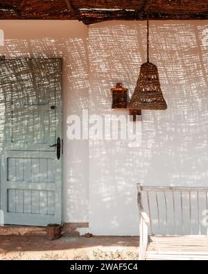 Eine Holzbank und eine Rattanlampe vor einer weißen Wand mit einem Eingang Stockfoto