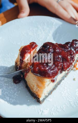 Draufsicht einer nicht erkennbaren Person, die mit einer Gabelscheibe Käsekuchen mit Himbeerauflage auf Porzellanplatte auf Holztisch schneidet, vertikales Bild für Stockfoto
