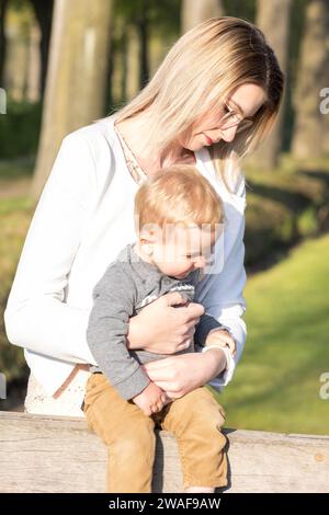 Dieses zarte Bild zeigt eine junge kaukasische Frau, die draußen sitzt und ein Kleinkind auf ihrem Schoß legt. Das Sonnenlicht filtert durch die Bäume und wirft ein warmes, flackerndes Licht über sie, das den weichen, fürsorglichen Ausdruck auf dem Gesicht der Frau hervorhebt, während sie auf das Kind herabblickt. Das Kind, gekleidet in einem grauen Hemd und einer braunen Hose, scheint sich in der behaglichen Umarmung wohl zu fühlen und verkörpert einen ruhigen Moment mütterlicher Zuneigung. Ruhige Momente in sonnenverwöhnter Gelassenheit. Hochwertige Fotos Stockfoto