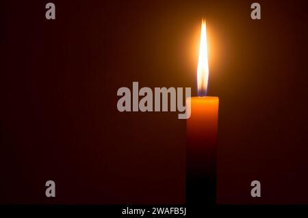 Eine einzelne brennende Kerzenflamme oder Licht leuchtet auf einer gelben Kerze, die auf dunkelrotem oder braunem Hintergrund auf dem Tisch in der Kirche zu Weihnachten, Beerdigung isoliert ist Stockfoto