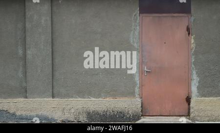 Braune schäbige Eisentür in einer grauen Betonwand eines unscheinbaren Hauses, leerer Minenraum an der Fassade eines alten Gebäudes neben einer geschlossenen Metalltür Stockfoto