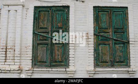 Zwei Fenster eines alten, weiß getünchten Hauses, verschraubt mit Holzläden mit abblätternder grüner Farbe, ein Fragment des Äußeren eines alten Gebäudes Stockfoto