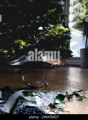 Weihnachtskugel aus zersplittertem Glas auf Wohnzimmerboden mit Weihnachtsbaum im Hintergrund Stockfoto