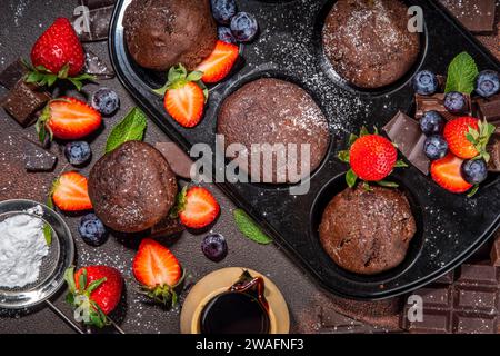 Schokoladen-Muffins mit Beeren. Muffin-Pfanne mit hausgemachten süßen, dunklen Schokoladen-Cupcakes mit Kakaosoße, Zuckerpulver und frischen Erdbeeren, Blaubeeren Stockfoto