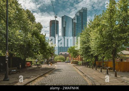 Eine von Bäumen gesäumte Kopfsteinpflasterstraße führt das Auge von der traditionellen bis zur zeitgenössischen Architektur der Wolkenkratzer von Manchester. Stockfoto