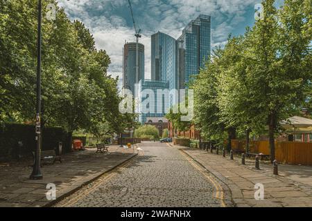 Eine von Bäumen gesäumte Kopfsteinpflasterstraße führt das Auge von der traditionellen bis zur zeitgenössischen Architektur der Wolkenkratzer von Manchester. Stockfoto