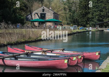 Kanus verzurrten sich auf dem Canal in Cromford, Derbyshire, England, und die Leute genossen Erfrischungen in einem Café im Hintergrund Stockfoto